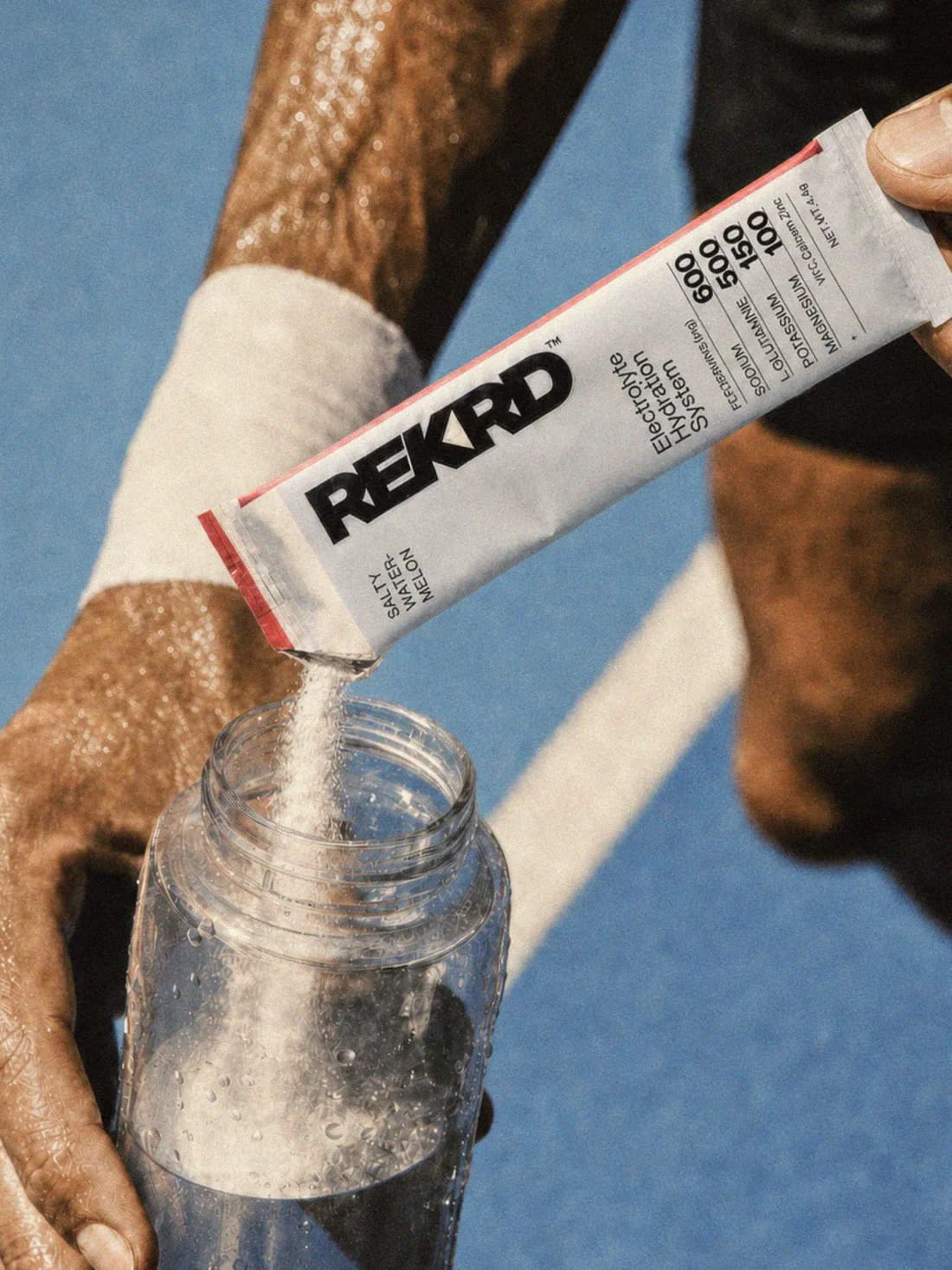 A REKRD Salty Watermelon sachet being poured into a clear water bottle on a tennis court